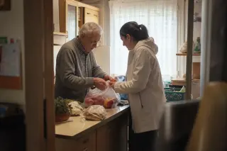 Senior man and young caregiver unpack groceries together in a softly lit, real home kitchen, viewed from the doorway, reflecting gentle collaboration and daily autonomy.