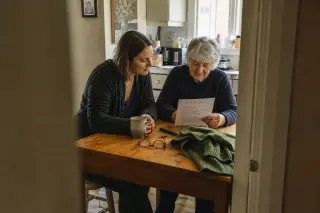 Senior woman and her adult daughter at a kitchen table, quietly reviewing a checklist together in gentle morning light, captured candidly from a doorway with a documentary feel.