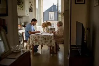 A senior woman and a male nurse share a quiet morning moment in a kitchen, sunlight streaming in, with subtle signs of daily life and care.