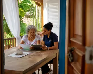 Elderly woman and a community worker collaborate at a sunlit wooden table on a Creole veranda, surrounded by tropical plants, in a natural and warm setting.